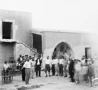 Men and boys gathered in a dusty yard at Panajiotis, Idalion, in front of a mud-brick building, several with arms raised.
