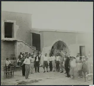 Farewell party at Panajiotis, Idalion: a crowd of men gathers in a dusty yard before a mud-brick building with an arched doorway