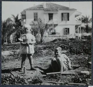 Two young children in a garden in Nicosia, one standing and one sitting by a small dug hole, with a white two-storey house behind.