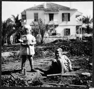 Two young children in a garden in Nicosia, one standing and one sitting in a shallow pit, with a white two-storey house behind