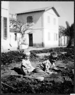 Two young children sitting on bare ground in front of a whitewashed two-storey house with shutters