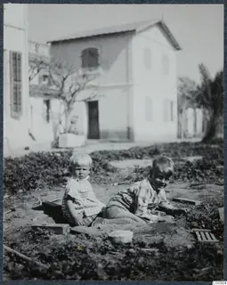 Two young children sitting on bare ground in front of a whitewashed two-storey building, Nicosia