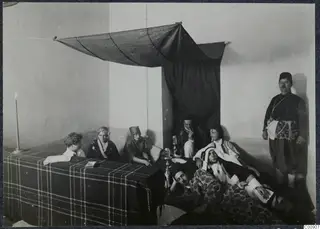 Staged "Oriental" party scene: people in dress-up clothes around a checked-covered bed under a draped dark cloth.