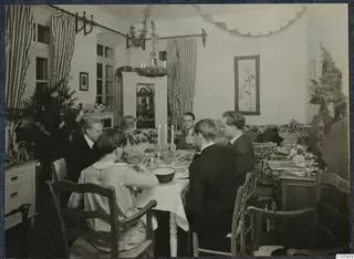 Christmas Eve 1927 meal: people seated at a candlelit table in a decorated room with striped curtains and a Christmas tree with flags
