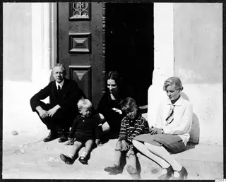 Five people sit on a whitened step in front of a wooden door, including two young children.