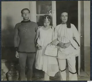 Three people in costume posing in a doorway at a masquerade, with large pottery vessels at the left edge.