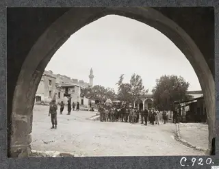 Kalavassos village square seen through a stone arch, with a minaret, trees and a group of men standing in front of an arcaded building