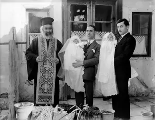 Priest and two men holding infants in white gowns outside a house, at the baptism of Giorkos, Nicosia