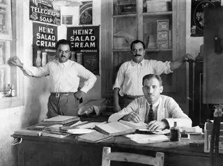 Three men in a small office with Heinz Salad Cream posters on the wall; one sits at a cluttered desk while two stand behind him.