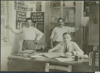 Office interior at Kalavasos with a seated man writing at a desk, two men behind him and a Heinz Salad Cream advertisement