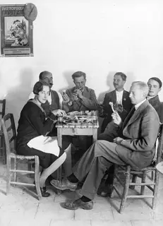 Six people seated at a small table in a whitewashed room, eating and drinking from small glasses.