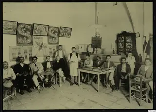 Group of about a dozen men and a waiter in a coffee house interior at Lapithos, seated and standing around tables.