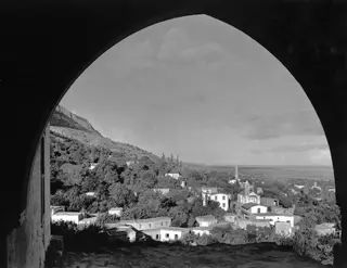 View through a pointed arch over a hillside village of white houses and trees, with a mountain slope on the left and a wide plain beyond.