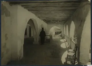 Priest in a dark robe walking along an arcaded gallery at Lambousa.