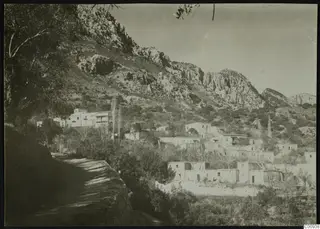 Village of white houses on a hillside below rocky mountains, with trees in the foreground and a track along a retaining wall.