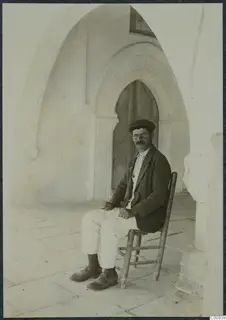 Solon seated on a wooden chair in an arched stone corridor, facing the camera.