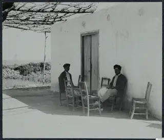 Two men sit on wooden chairs outside a whitewashed building at Karavas, beside a wooden door and a vine pergola.