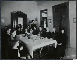 Group of people seated and standing around a dinner table in a whitewashed room with a carved wooden cupboard.