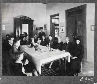 Group of about fifteen people seated and standing around a dinner table with a white cloth in a plastered room