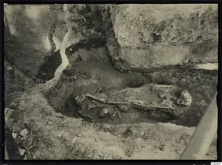 Skeleton lying in an excavated trench at Lapithos, with bones exposed in dark soil beside a large rock face.