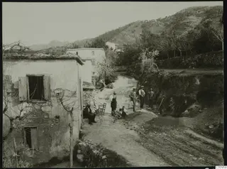 Village lane at Kylistra, Lapithos, with a rendered stone house and several people standing and sitting on a dirt road