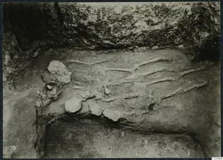 Skeleton lying in an excavated pit at Idalion, with small vessels beside it and a rock face above