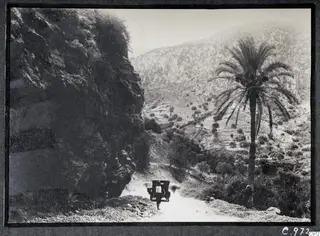 Road from Kyrenia to Nicosia winding past a rock face, a palm tree and an old car with a man beside it