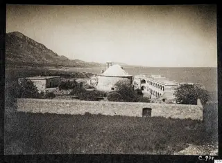 Monastery at Lambousa seen from the west, with a low stone wall in front, bushes between the buildings, and hills and sea behind
