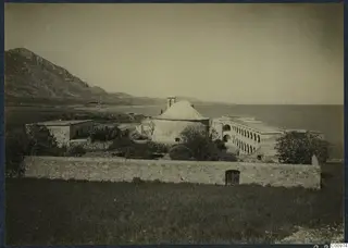 Monastery buildings at Lambousa seen from a walled field, with a domed round structure and arcaded range beside the sea.