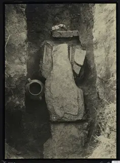 Tomb 422 at Lapithos Kastros during excavation, with a large stone slab, a handled jug and sherds in the cut.