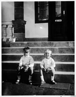 Two young boys sit side by side on a stone staircase in front of a dark wooden door.