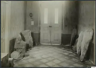 Interior of a room with several large stone sculptures leaning against the walls and lying on the tiled floor.