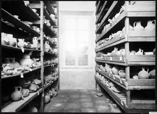 Storage room with wooden shelves holding many pottery vessels, many with paper tags, and a tall window at the far end.