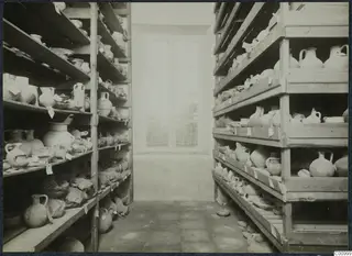 Storage room with wooden shelves on both sides filled with pottery jugs and bowls, many with paper tags, and a window at the back.