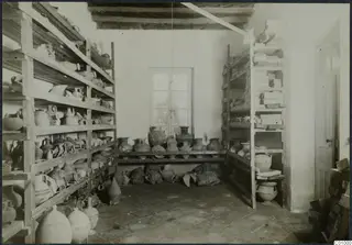 Storage room with open wooden shelves filled with pottery jugs and jars, and larger jars standing on a low shelf below a window.