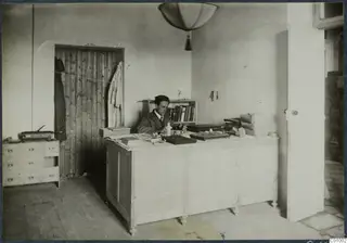Einar Gjerstad seated at a large wooden desk in an office room, with a bookshelf and a door beside him.