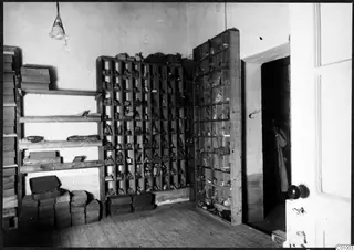 Storeroom with wooden shelves and open pigeonhole racks holding small objects, with a person in a doorway at right.
