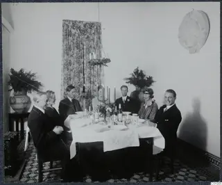 Christmas Eve 1928 dinner: seven people seated at a table with a candelabra, bottles and a white cloth.