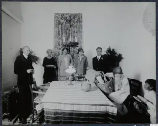 Christmas Eve 1928: adults stand in a room while a figure in fur sits at a table with a basket and parcels; a child sits at right.