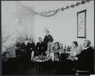 Seven people in a decorated living room on Christmas Eve, with a fruit basket and a lit candle on a low table.