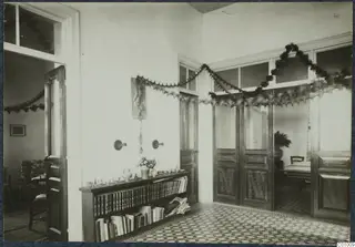 Interior of a room with a tiled floor, a wooden bookcase of books and vessels, and garlands of greenery over the doors.