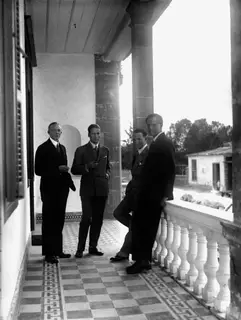 Four men in suits standing on a tiled veranda with a white balustrade, looking toward the camera.