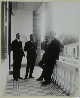 Four men in dark suits standing on a tiled veranda beside a stone column and white balustrade, Nicosia.