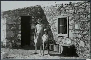Woman and a young boy standing outside a stone house at Vouni, with a doorway and a window.