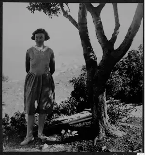 Woman in a long dress standing beside a tree on a hillside overlooking the sea