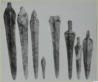 Row of about eight bronze blades and tanged tools laid upright on a plain grey background