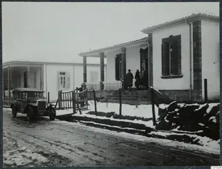 The Swedish house in Nicosia, a white building with a columned veranda, people on its steps and an old car in snow.