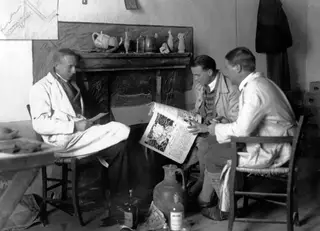 Three men in a room by a fireplace; the man in the middle holds open a printed newspaper, Nicosia