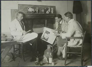 Three men seated in a room by a fireplace, one reading a newspaper, with bottles and a large jug on the floor.