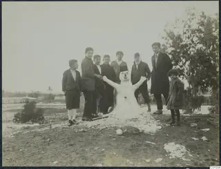Eight children and a young man standing around a small snowman on bare ground, Nicosia